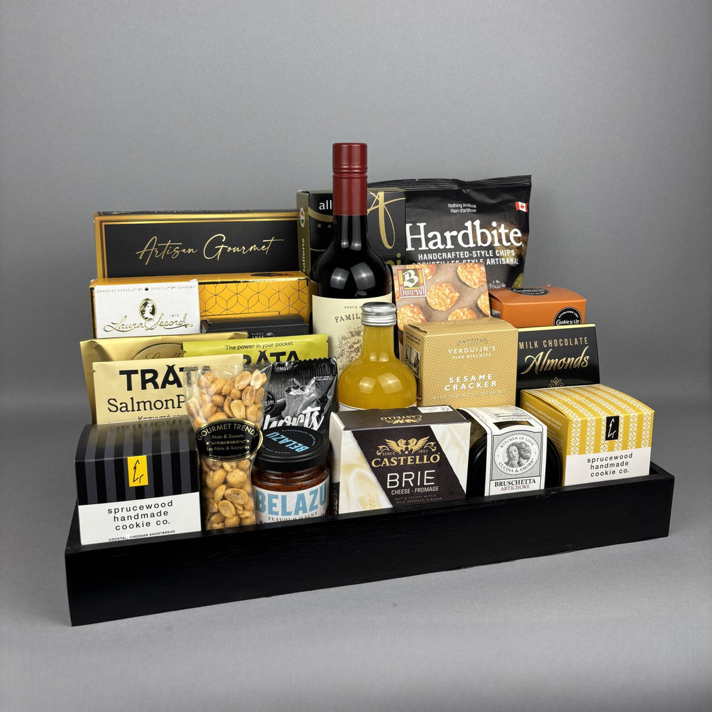 A gourmet gift hamper arranged on a black rectangular tray. The tray holds assorted packaged food items including a bottle of red wine, a bottle of vinegar, crackers, nuts, cheese, chocolates, and cookies from brands such as Laura Secord, Hardbite, Hamlet, Belazu, and Castello.