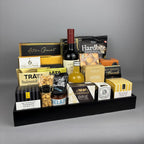 A gourmet gift hamper arranged on a black rectangular tray. The tray holds assorted packaged food items including a bottle of red wine, a bottle of vinegar, crackers, nuts, cheese, chocolates, and cookies from brands such as Laura Secord, Hardbite, Hamlet, Belazu, and Castello.