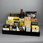 A gourmet gift hamper arranged on a black tray. The tray contains a bottle of red wine, a bottle of vinegar, assorted boxed cheeses, crackers, nuts, chocolates, and packaged snacks.