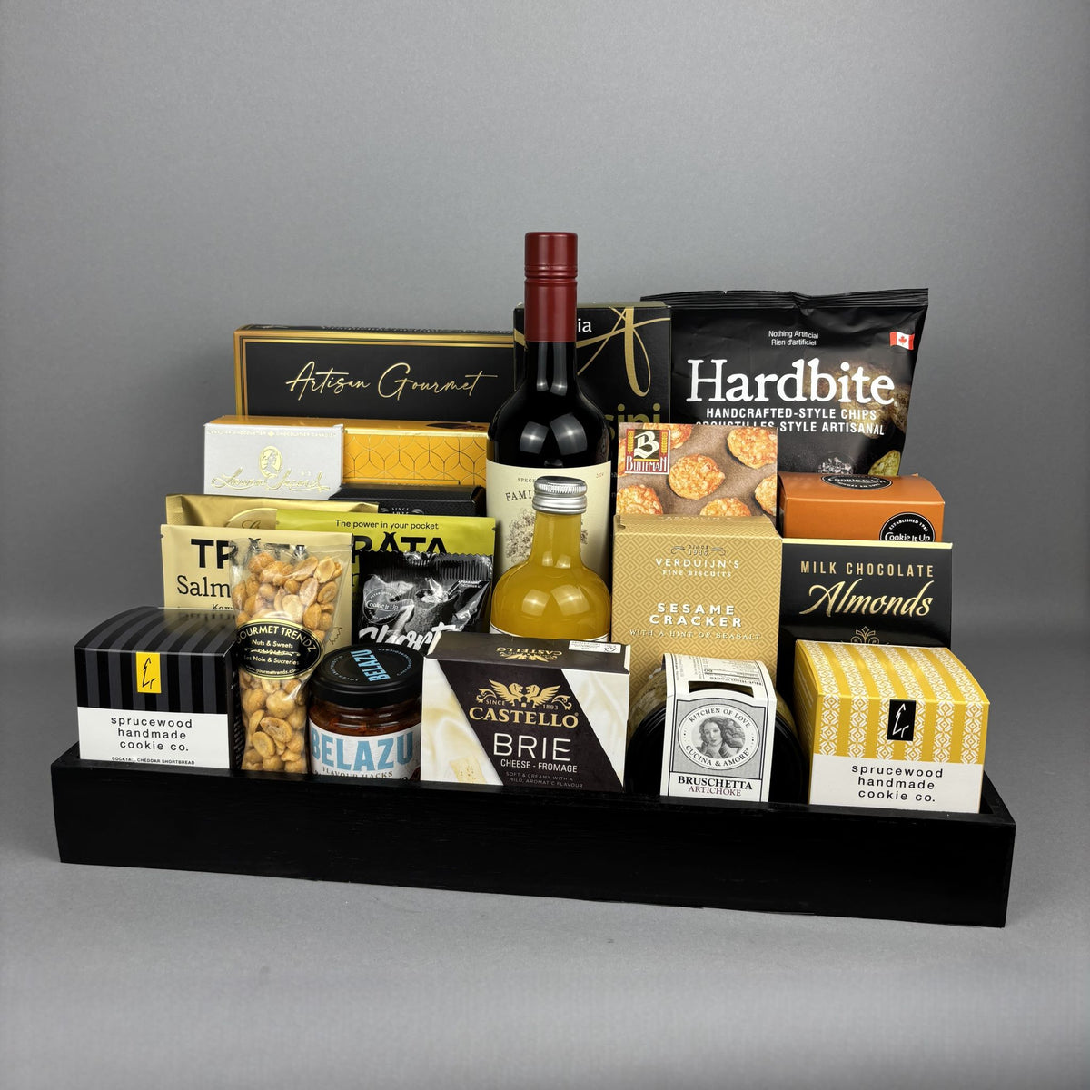A gourmet gift hamper arranged on a black tray. The tray contains a bottle of red wine, a bottle of vinegar, assorted boxed cheeses, crackers, nuts, chocolates, and packaged snacks.