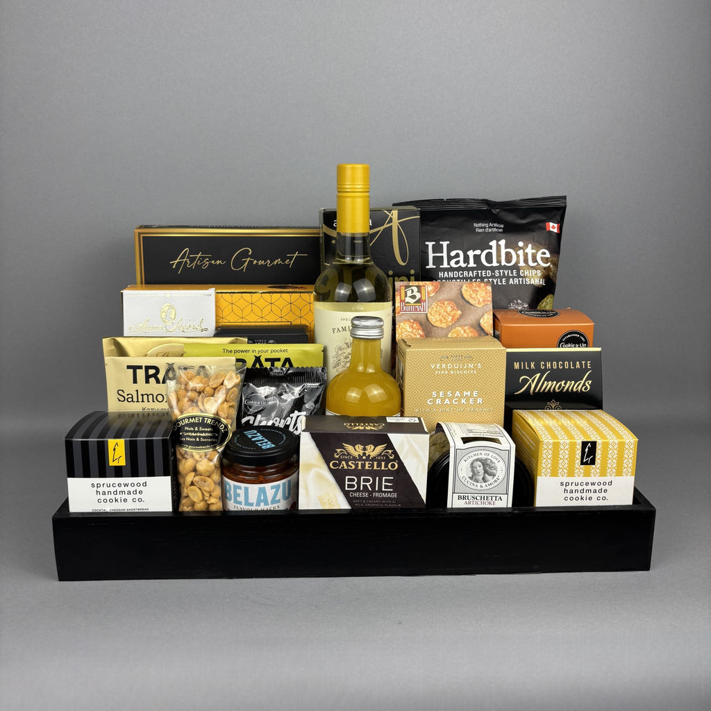 A gourmet gift hamper arranged on a black tray, featuring packaged snacks, chocolates, nuts, a bottle of vinegar, a bottle of white wine, and jars of bruschetta and spreads.