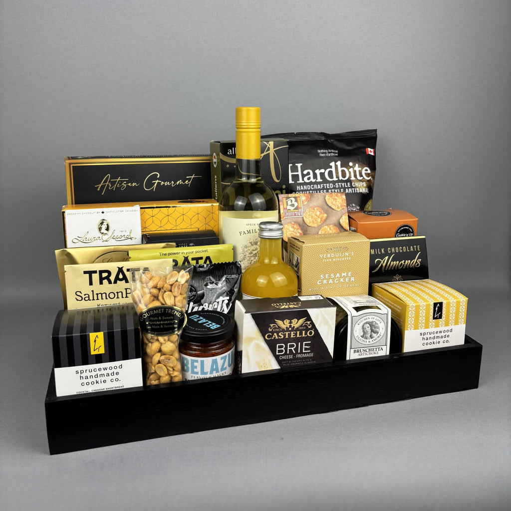 A curated gourmet gift hamper arranged on a black rectangular tray. The assortment includes packaged snacks, crackers, a bottle of vinegar, a bottle of white wine, and boxed sweets and cheeses.