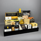 A curated gourmet gift hamper arranged on a black rectangular tray. The assortment includes packaged snacks, crackers, a bottle of vinegar, a bottle of white wine, and boxed sweets and cheeses.