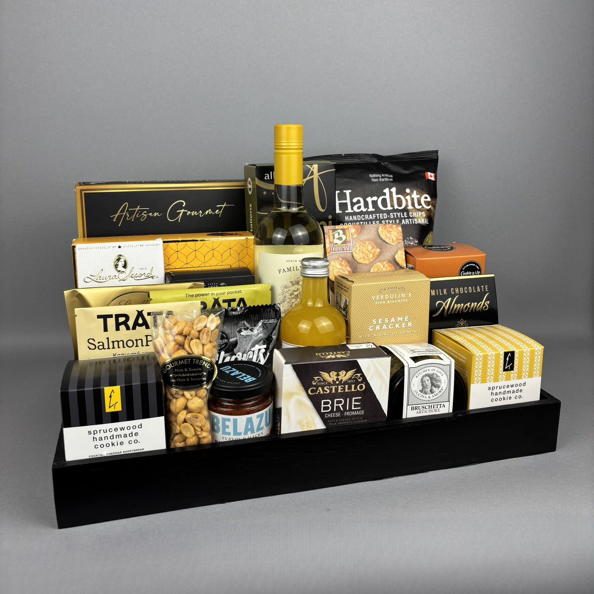 A curated gourmet gift hamper arranged on a black rectangular tray. The assortment includes packaged snacks, crackers, a bottle of vinegar, a bottle of white wine, and boxed sweets and cheeses.