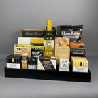 A curated gourmet gift hamper arranged on a black tray. The assortment includes cheese, crackers, nuts, chocolate, and a bottle of vinegar, with visible brands such as Castello Brie, Hardbite, Hamlet, and Laura Secord.