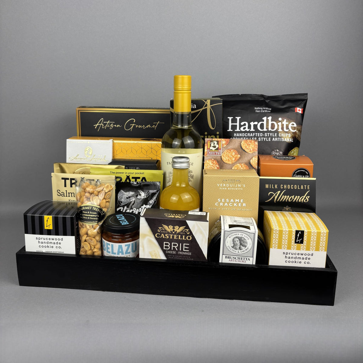 A curated gourmet gift hamper arranged on a black tray. The assortment includes cheese, crackers, nuts, chocolate, and a bottle of vinegar, with visible brands such as Castello Brie, Hardbite, Hamlet, and Laura Secord.