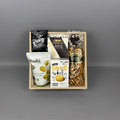 A gourmet gift box containing various snacks and cheeses, including Poshi Green Olives, Castello Brie Cheese, Canadian cheddar biscuits, pretzels, and chocolate covered oreo, arranged in a light wood tray.