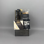 A gift basket arrangement of gourmet snacks and cheeses displayed on a dark wooden base The assortment includes Castello Brie cheese, Black Pepper Crackers, biscotti, pretzels, and a milk chocolate shortbread cookie.