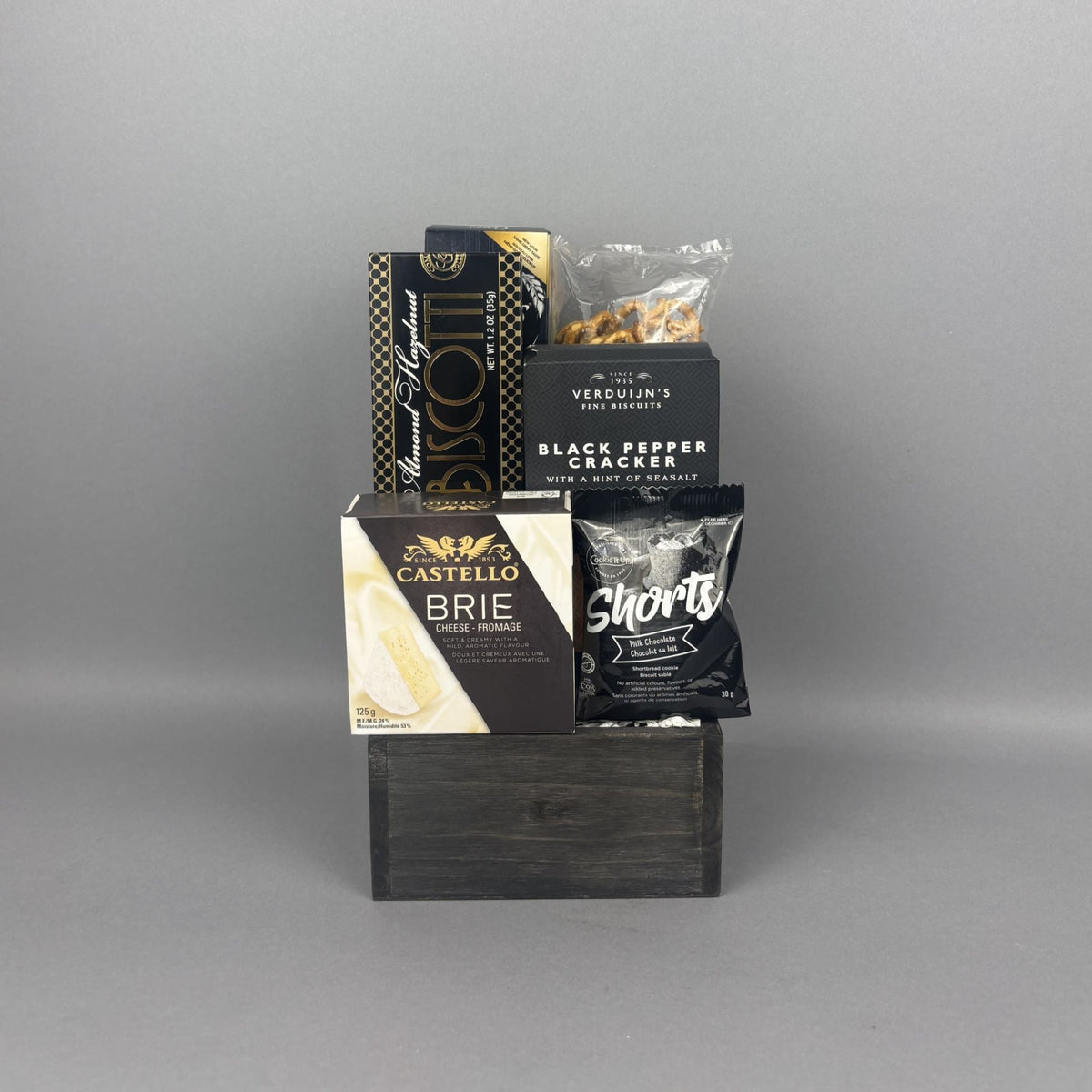 A gift basket arrangement of gourmet snacks and cheeses displayed on a dark wooden base The assortment includes Castello Brie cheese, Black Pepper Crackers, biscotti, pretzels, and a milk chocolate shortbread cookie.