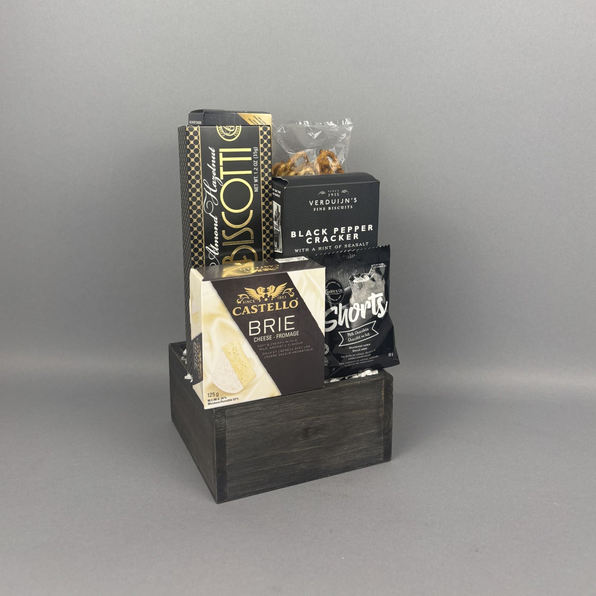A gourmet gift hamper in a dark wooden box, featuring a box of almond hazelnut biscotti, a box of Castello Brie cheese, a box of Verduijn's Black Pepper wafers, and a bag of milk chocolate shortbread cookies.