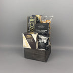 A gift basket arrangement in a dark wooden crate containing packaged snacks and cheeses, including a box of Castello Brie, a box of Verduijn's Black Pepper Crackers, and other packaged items.