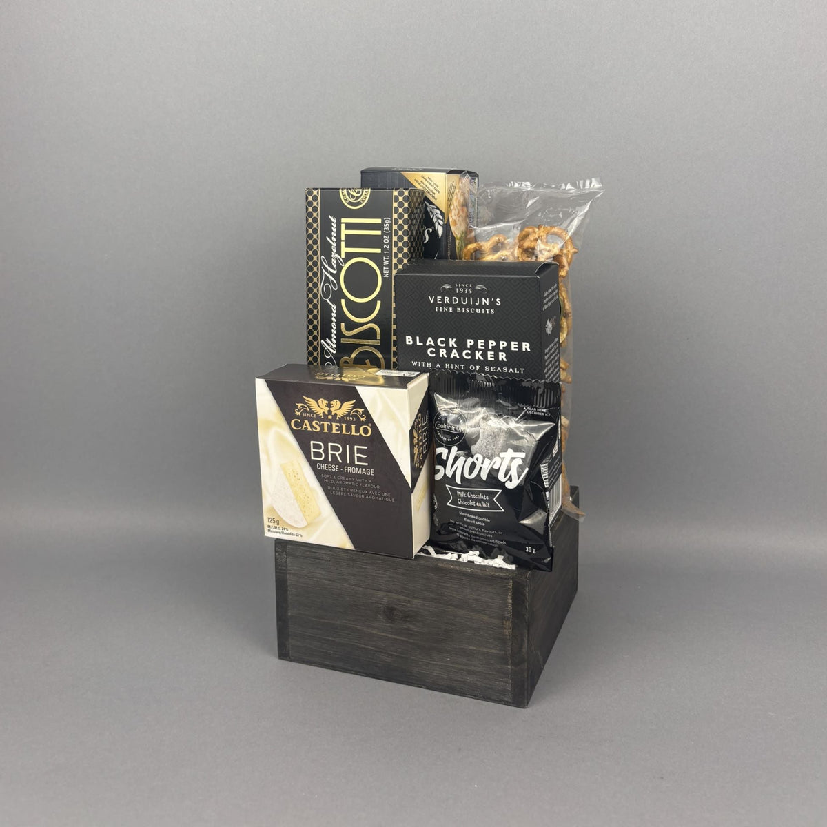 A gift basket arrangement in a dark wooden crate containing packaged snacks and cheeses, including a box of Castello Brie, a box of Verduijn's Black Pepper Crackers, and other packaged items.