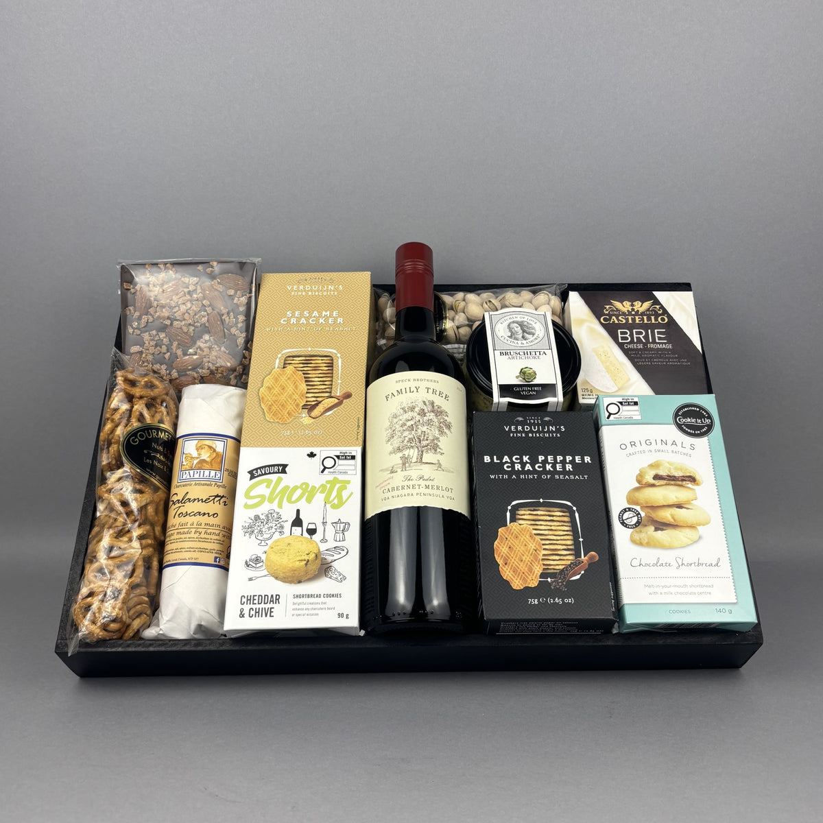 A gourmet gift hamper arranged in a black tray, featuring a bottle of Family Tree Red Wine, Castello Brie cheese, Shorts cheddar & chive biscuits, Black Pepper crackers, assorted nuts and pretzels, chocolates, and artichoke bruschetta.
