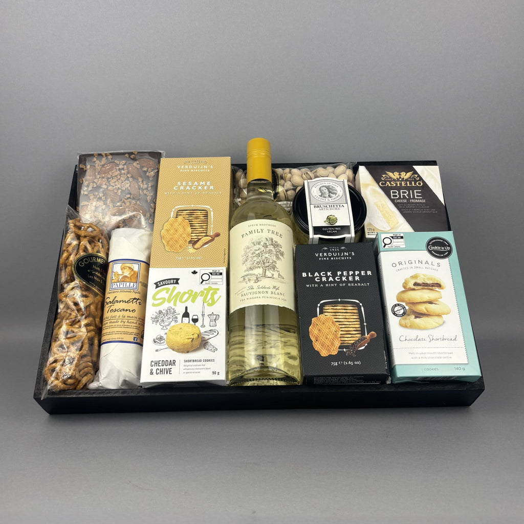 A gourmet gift hamper arranged in a black tray, featuring a bottle of white wine, cheese, crackers, pretzels, chocolates, and packaged snacks.