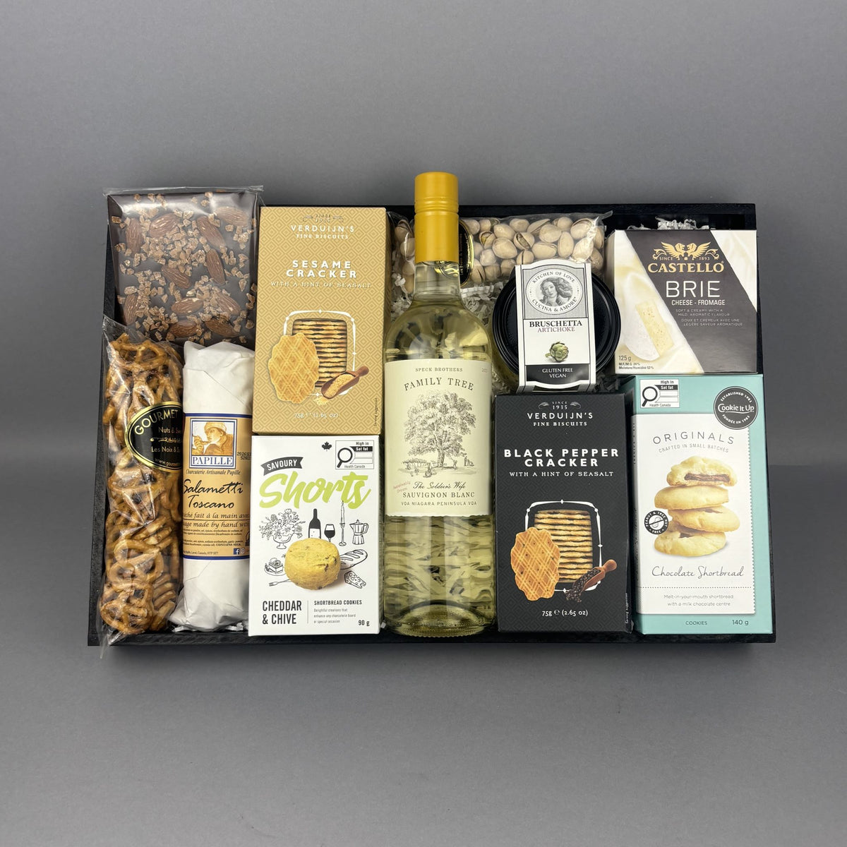 A curated gourmet gift box containing a bottle of Family Tree Sauvignon Blanc, Castello Brie cheese, Pretzels, cheddar & chive biscuits, Black Pepper Cracker, and other snacks arranged in a black tray.