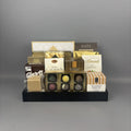A gourmet chocolate and snack gift basket arranged on a black tray. The assortment includes milk chocolate almonds, shortbread cookies, caramel candies, and various boxed chocolates from brands like Made Chocolate and Andea Chocolates.