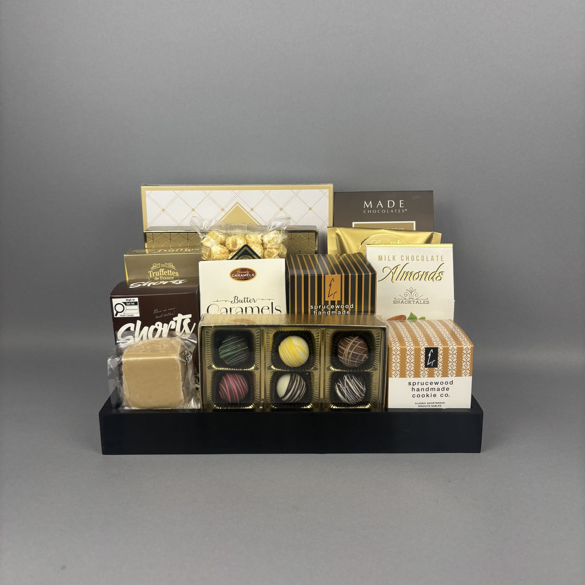A gourmet chocolate and snack gift basket arranged on a black tray. The assortment includes milk chocolate almonds, shortbread cookies, caramel candies, and various boxed chocolates from brands like Made Chocolate and Andea Chocolates.