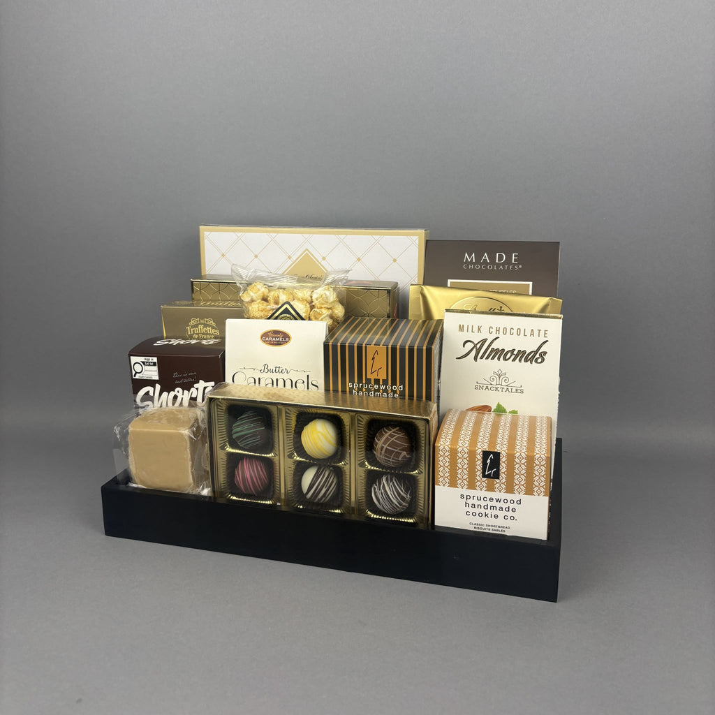 A gourmet chocolate and almond gift basket arranged on a black tray. The assortment includes various boxed chocolates, pralines, almonds, and a bag of cookies, with several brands including Lindt, Sprucewood, Mad Chocolates, Aunt Gloria's and more.