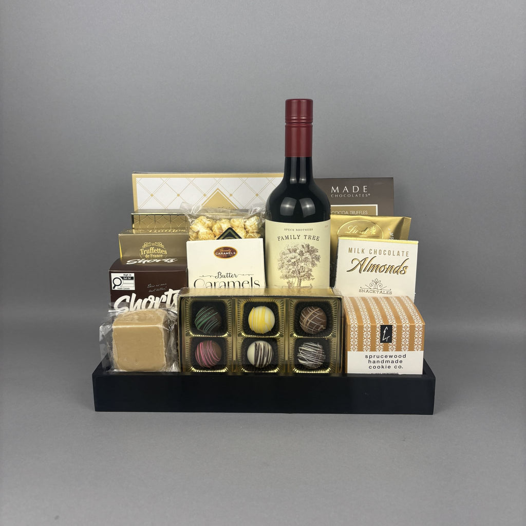 A gourmet gift hamper arranged on a black tray, featuring a bottle of red wine, assorted chocolates, milk chocolate almonds, caramel candies, and boxed treats.