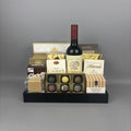 A gourmet gift hamper arranged on a black tray, featuring a bottle of red wine, assorted chocolates, milk chocolate almonds, caramel candies, and boxed treats.