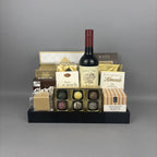 A gourmet gift hamper arranged on a black tray, featuring a bottle of red wine, assorted chocolates, milk chocolate almonds, caramel candies, and boxed treats.