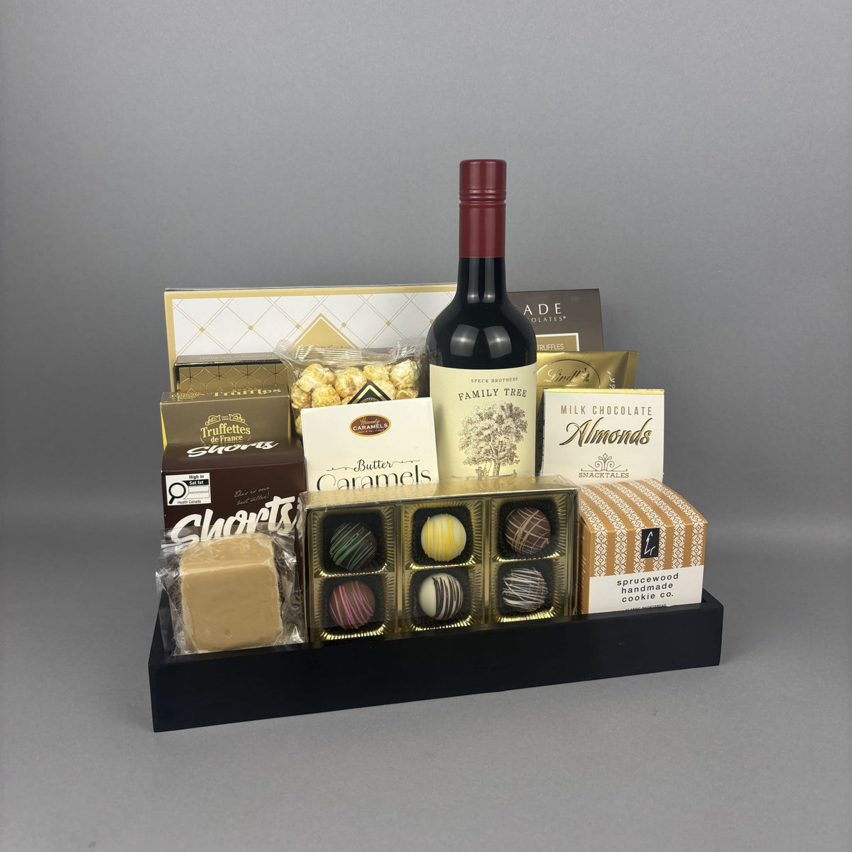 A gourmet gift hamper featuring a bottle of red wine, assorted chocolates, caramels, almonds, and cheese, arranged  on a black serving tray.