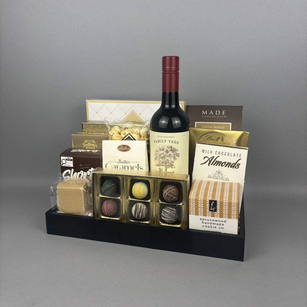 A gourmet gift hamper arranged on a black tray, featuring a bottle of red wine, assorted chocolates, butter caramels, milk chocolate almonds, and other packaged treats.