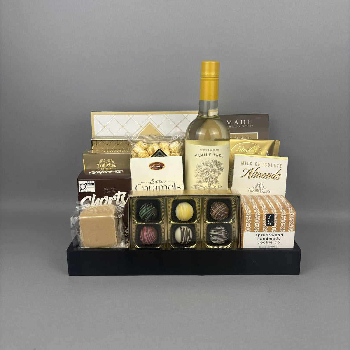 A gourmet gift hamper featuring an assortment of chocolates, a bottle of white wine, and various boxed treats arranged on a black tray.