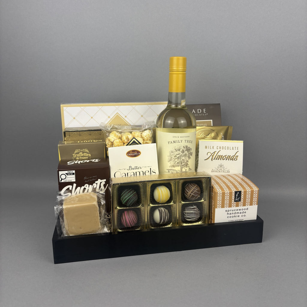 A gourmet gift hamper arranged on a black tray, featuring a bottle of white wine, assorted chocolates, caramels, almonds, and cookies.