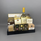 A gourmet gift hamper arranged on a black tray, featuring a bottle of white wine, assorted chocolates, caramels, almonds, and cookies.