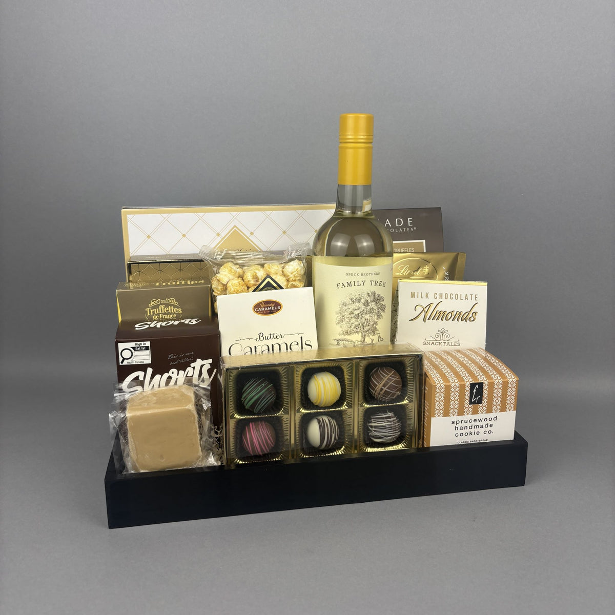 A gourmet gift hamper arranged on a black tray, featuring a bottle of white wine, assorted chocolates, caramels, almonds, and cookies.