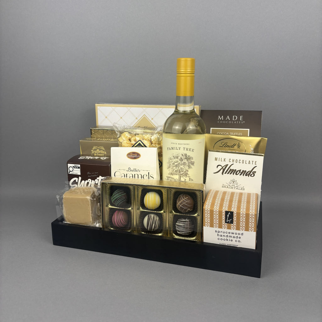 A gourmet gift hamper arranged on a black tray, featuring a bottle of white wine, assorted chocolates, almonds, caramels, and packaged snacks.
