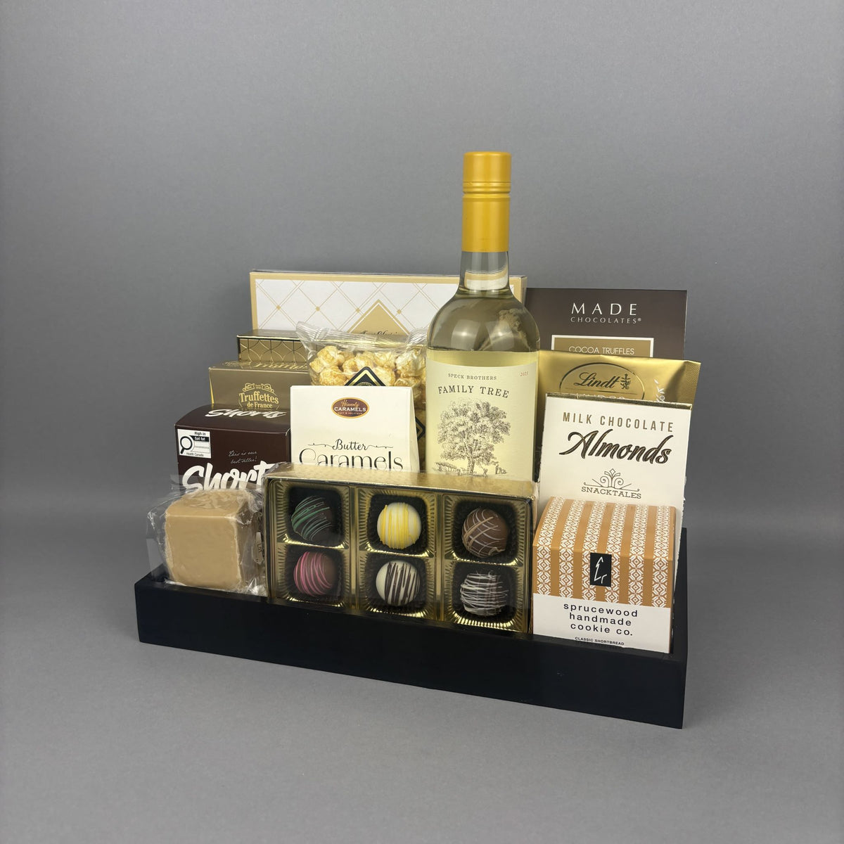 A gourmet gift hamper arranged on a black tray, featuring a bottle of white wine, assorted chocolates, almonds, caramels, and packaged snacks.