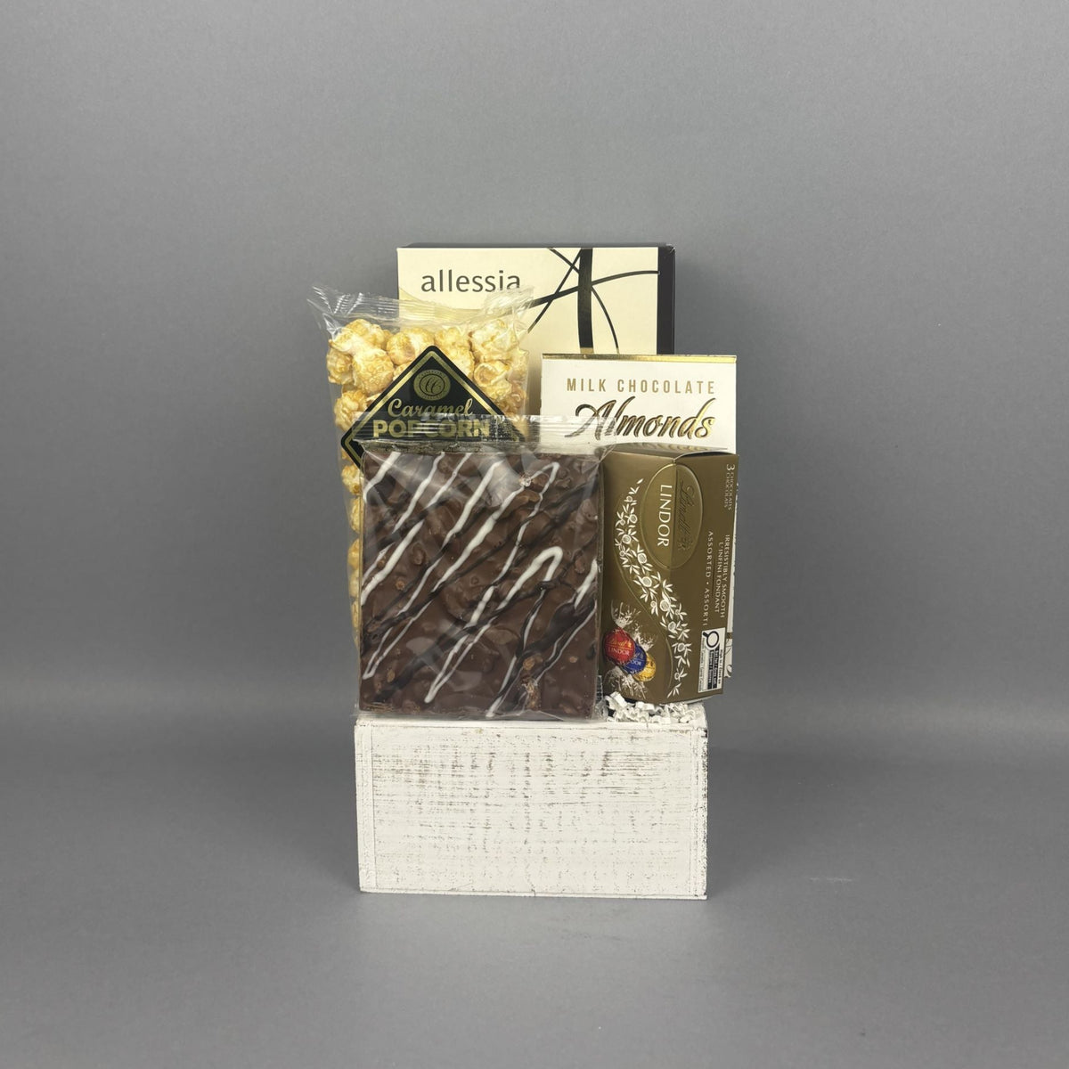 A gift basket of assorted chocolates and snacks displayed on a white wood basket. The assortment includes a milk chocolate almonds, a chocolate bark bar, a bag of caramel popcorn, a box of Allessia biscotti, and Lindt truffles.