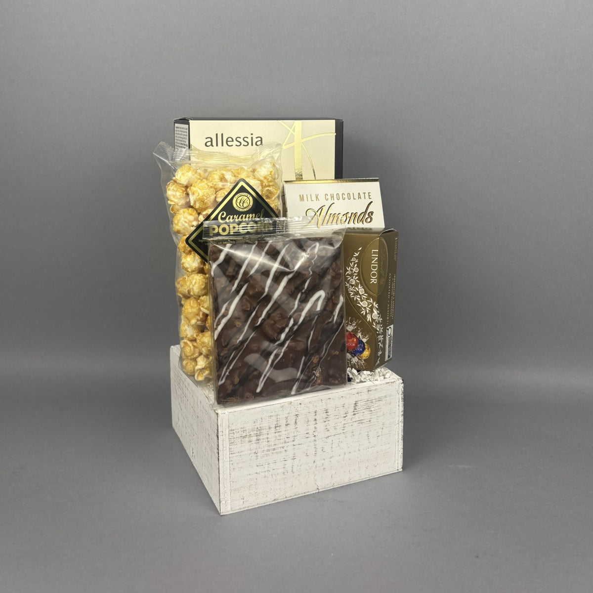 A gift basket arrangement of snacks and chocolates displayed on a white wooden basket. The assortment includes Allessia biscotti, a box of chocolate-covered almonds, and a box of Lindt chocolate truffles.