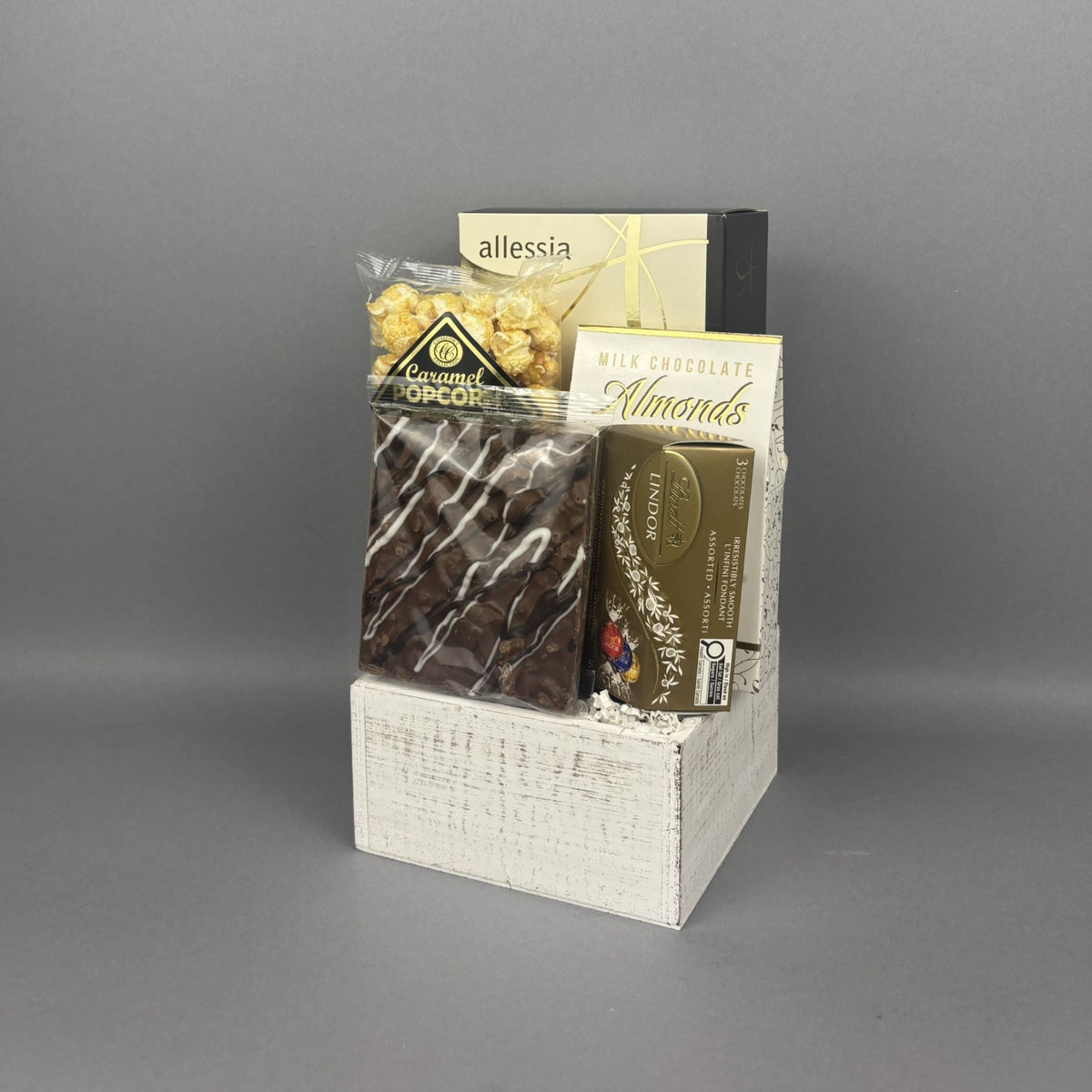 A gourmet chocolate and almond gift basket arranged in a white wood basket. The basket contains a box of Allessia biscotti, a bag of caramel popcorn, chocolate-covered almonds, a box of Lindt truffles, and a milk chocolate bark bar.